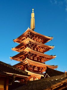 View larger photo: A vibrant image of a traditional Japanese pagoda with intricate architectural details, illuminated by the warm glow of the setting sun. The pagoda stands tall against a clear, deep blue sky, highlighting its golden spire and tiered roofs adorned with delicate eaves.