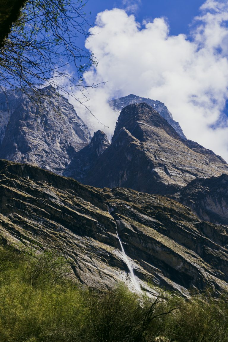 A towering mountain range under a partly cloudy blue sky, with a waterfall cascading down the rocky slopes, and greenery in the foreground.