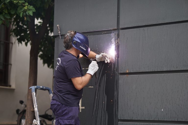 A worker wearing a protective helmet and gloves is welding a metal structure outdoors. There is a stepladder leaning against the wall and a tree in the background. Sparks are visible at the point of welding.