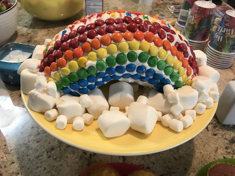 M&M decorated birthday cake in a rainbow arch with marshmallows surrounding them like clouds