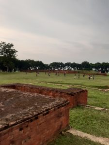 Vasu Bihar is one of the famous archaeological sites in Bangladesh. A group of people playing soccer on a grassy field surrounded by trees and ancient brick structures.