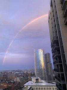 View of a Boston cityscape featuring modern high-rise buildings with a prominent curved reflective glass tower. A vibrant rainbow stretches across the sky, creating an arch above the buildings.