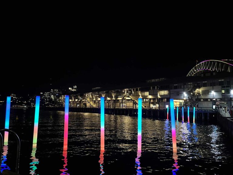 A nighttime waterfront scene featuring a series of colorful, rainbow light illuminated vertical poles reflecting on the water.