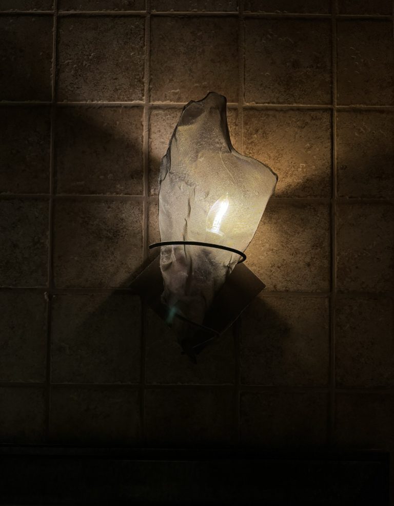 A partially translucent lighting sconce in a dark area against a brownish-red tile wall.