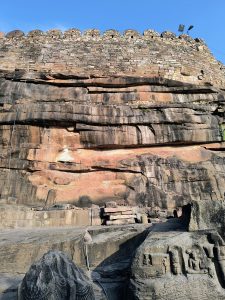 Ancient stone carvings and rugged rock faces form the base of Kalinjar Fort's formidable walls under a clear blue sky.