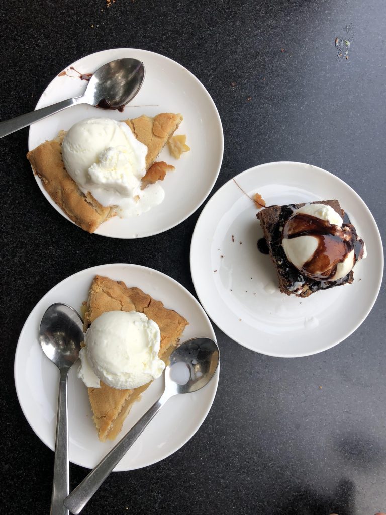 Three white plates hold desserts: the top left and bottom plates have pie slices with vanilla ice cream and spoons, while the right plate has chocolate cake with vanilla ice cream and chocolate syrup.