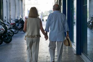 View larger photo: Senior couple walks hand in hand down a tiled sidewalk, with motorcycles parked along the side. The man holds a hat in his free hand.