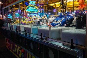 A brightly lit arcade claw machine filled with various colorful stuffed animals. The machine's control panel is visible in the foreground, while neon lights and other arcade machines can be seen in the background, creating a lively and vibrant atmosphere.