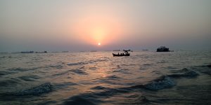 A serene seascape at sunset with a calm sea and gentle waves in the foreground. A small boat with people is silhouetted against the setting sun, while several large ships are scattered along the horizon.