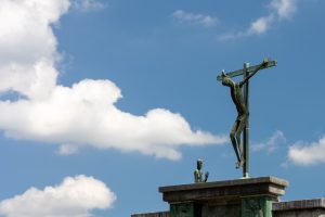 A statue of a man on a cross along with a man kneeling to his side against a background of a blue sky with a few clouds.