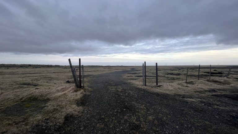 Dirt and gravel road winds between a wood and wire fence along Tjóevegur leading up to Fossálar Waterfall (Iceland)