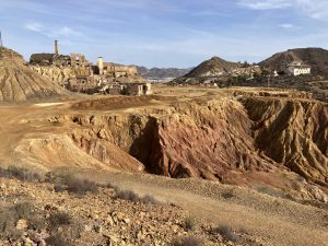 A barren landscape featuring erosion-scarred, reddish-brown terrain with rugged hills and derelict industrial buildings, including structures with tall chimneys, in the distance. Sparse vegetation dots the arid ground, and a clear blue sky stretches overhead