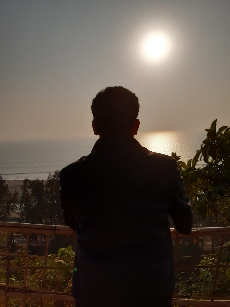 A person with short hair stands facing the sunlit ocean, surrounded by greenery and a railing in the foreground