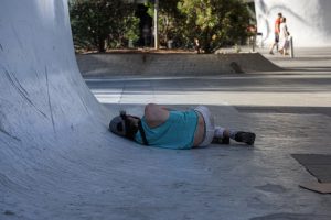 A person is lying on their side, leaning against a curved concrete surface. The background shows some trees and a few pedestrians walking.