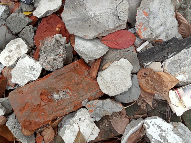 A close-up of various chunks of broken concrete, brick pieces, and dried leaves. Among the rubble, different textures and colors of the debris are visible, including gray and white concrete fragments, red brick, and brown autumn leaves.