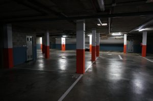 A dimly lit underground parking garage with several empty parking spaces. Concrete columns, some marked with orange paint, are spaced throughout the area. A door and a fire extinguisher are visible on the walls.