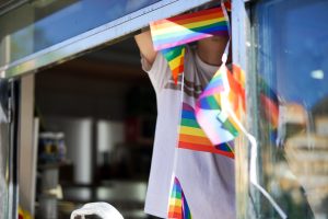 A shopkeeper is hanging rainbow flags on a window, with the interior of a store visible in the background.