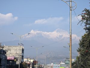 View of a city street with buildings, streetlights, and power lines, leading to a backdrop of snow-capped mountains of Annapurna Range, Himalay.