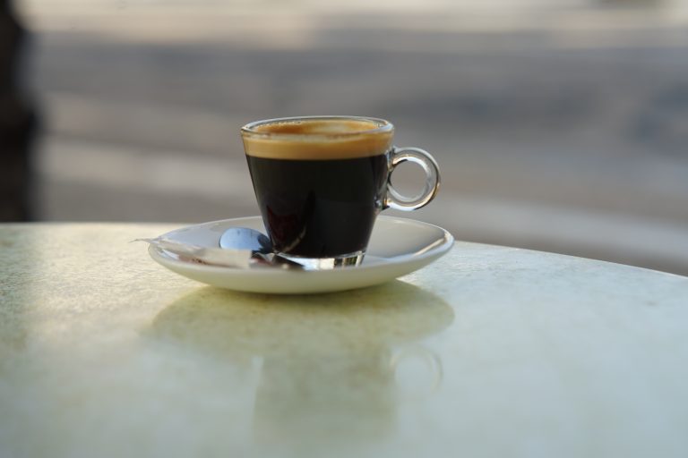 A small cup of espresso coffee, is placed on a white saucer with a spoon on a light-colored table.