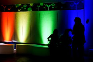 View larger photo: Three silhouetted people are interacting in front of a brightly lit wall. The wall is illuminated with colors fading from red to green to blue, creating a gradient effect resembling a rainbow.