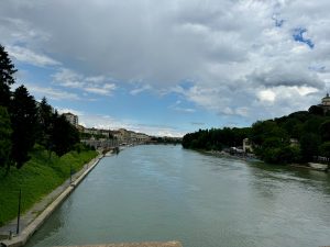 这张照片拍摄了意大利都灵波河（Po River）的宁静景象，摄于穿越翁贝托大桥（Umberto Bridge）后。河水平静地流着，两岸绿树葱郁。在左岸，一条步行小路与河流平行，两旁是树木和灯柱。远处可以看到建筑物和房屋，还有一些小船