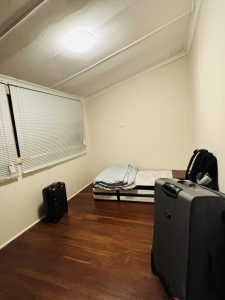 A small, minimally furnished bedroom with a single mattress on the floor, covered with a folded blanket. The room has white walls and a wooden floor.