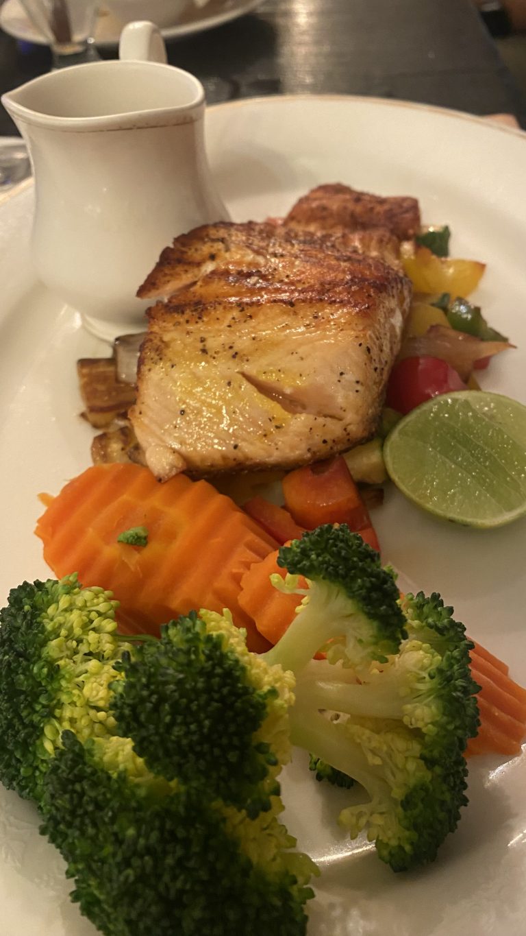 Grilled salmon fillet served with sautéed vegetables, including sliced bell peppers, carrots, and broccoli, accompanied by a slice of lime and a small white pitcher.