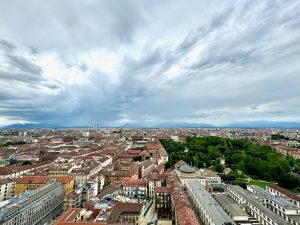  An expansive view of Torino, Italy, taken from the top of the Cinema Museum. The photo showcases a wide cityscape with a mix of historic and modern buildings, red-tiled roofs, and lush green parks. The horizon is lined with distant mountains under a dramatic sky filled with clouds, giving a sense of depth and scale to the city below.