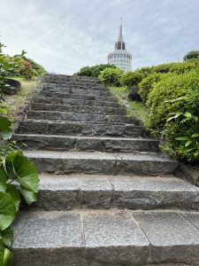 A stone staircase leads upwards through a garden with green shrubs and small flowering bushes on either side. In the background, partially obscured by greenery, a tall, modern building with a pointed spire is visible under a cloudy sky.