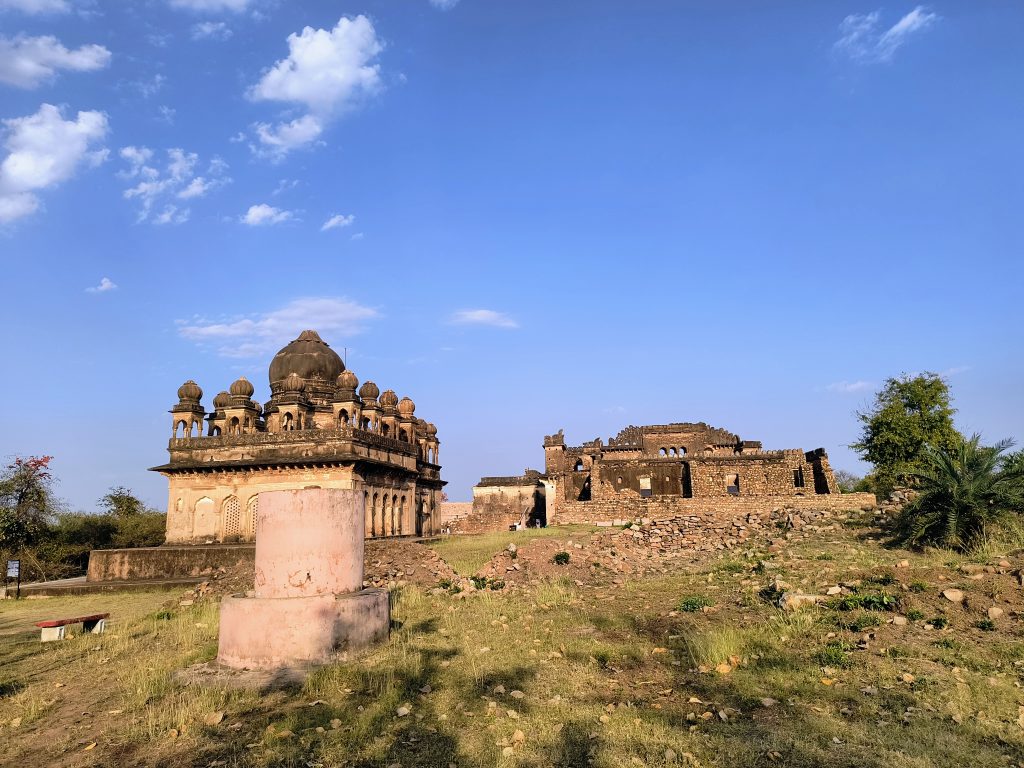 Historic domed structures and ruins within Kalinjar Fort, set against a bright blue sky with scatter...