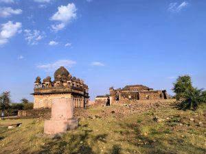 Historic domed structures and ruins within Kalinjar Fort, set against a bright blue sky with scattered clouds.
