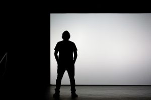 View larger photo: A person standing alone in front of a large, bright white screen, with their silhouette visible against the light background.