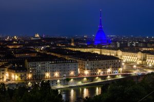 A night time view of the Italian city of Torino.  There is a river in the foreground with the city laid out stretching to the horizon.  The building and street lights are most prominent on a main thoroughfare which leads to the landmark building, Mole Antonelliana, which is illuminated a shade of blue.