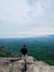 View larger photo: A person stands on a rocky cliff overlooking a vast, green forested landscape, with mountains and a cloudy sky in the distance.