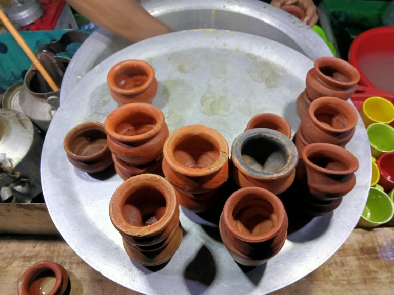 A metal tray holds several small, empty clay cups. On the left, a stainless steel kettle is partially visible, and to the right, there are colorful small plastic bowls. A person’s hand is visible in the background, holding another clay cup.