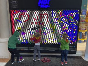 View larger photo: Three children designing a rainbow design on an oversized multicolor light pegboard