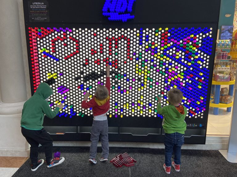 Three children designing a rainbow design on an oversized multicolor light pegboard