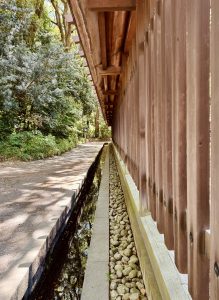 A perspective view of a wooden structure, possibly a traditional Japanese fence or wall, with a small stone-filled drainage ditch running parallel to it. The scene is set in a natural, outdoor environment with trees and foliage on the left side, creating a serene and peaceful atmosphere. 