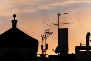 View larger photo: Silhouette of a rooftop with various antennas and satellite dishes against a dusk sky.