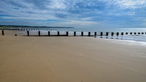 A sandy beach extends from the left of the frame to a diagonal near the right edge, the tide has recently gone out and the sand is smooth and wet. The beach features wave breaks extending into the sea. It is a cloudy day but quite bright.