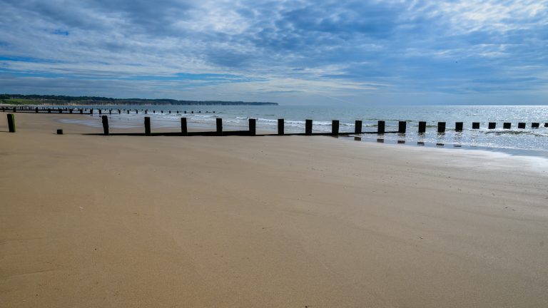 A sandy beach extends from the left of the frame to a diagonal near the right edge, the tide has recently gone out and the sand is smooth and wet. The beach features wave breaks extending into the sea. It is a cloudy day but quite bright.