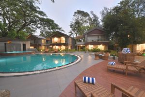 View larger photo: The image shows a swimming pool at a resort. The pool is surrounded by outdoor furniture, trees, and buildings. The sky is visible in the background.