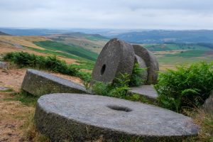 A landscape view of the Peak District in the UK. The foreground is dominated by large stone disks with a green patchwork of rolling hills extending to the horizon. It is a cloudy day with no shadows.