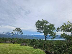 View larger photo: A scenic landscape featuring lush greenery with trees in the foreground and a distant view of mountains partially covered by clouds. The sky is mostly overcast with some patches of blue. 