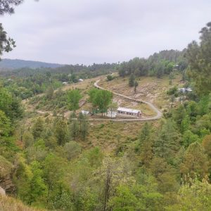 A hilly landscape with a winding road leading to several small buildings surrounded by trees. The area is lush with greenery and vegetation, with a mixture of dense forested sections and open grassy areas. The sky is overcast.