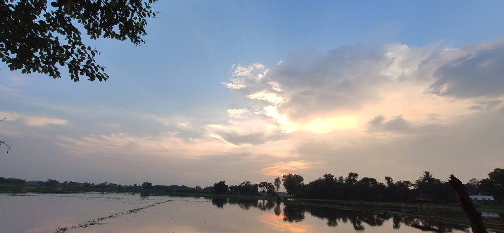 A serene evening sky with a mix of blue and white clouds, as the sun begins to set. The horizon features silhouetted trees, and the sky is reflected in the calm water of a nearby field.