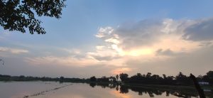  A serene evening sky with a mix of blue and white clouds, as the sun begins to set. The horizon features silhouetted trees, and the sky is reflected in the calm water of a nearby field.
