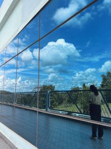 A person standing near a railing, reflected in large windows of a building. The windows also reflect a bright blue sky filled with fluffy white clouds and some green trees in the background. The person is wearing a beige shirt and dark pants.