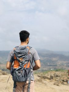 A person with short dark hair, carrying an orange and grey bag, is observing a scenic mountainous landscape.