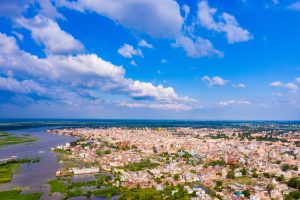 View larger photo: Beauty of Yamuna Ji, Vrindavan. Overview of a town with water on one edge, blue sky with light clouds above.